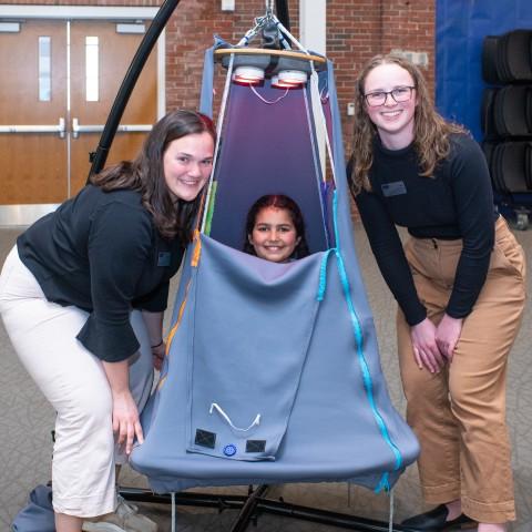 A child sits inside a suspended tent and two students pose around her